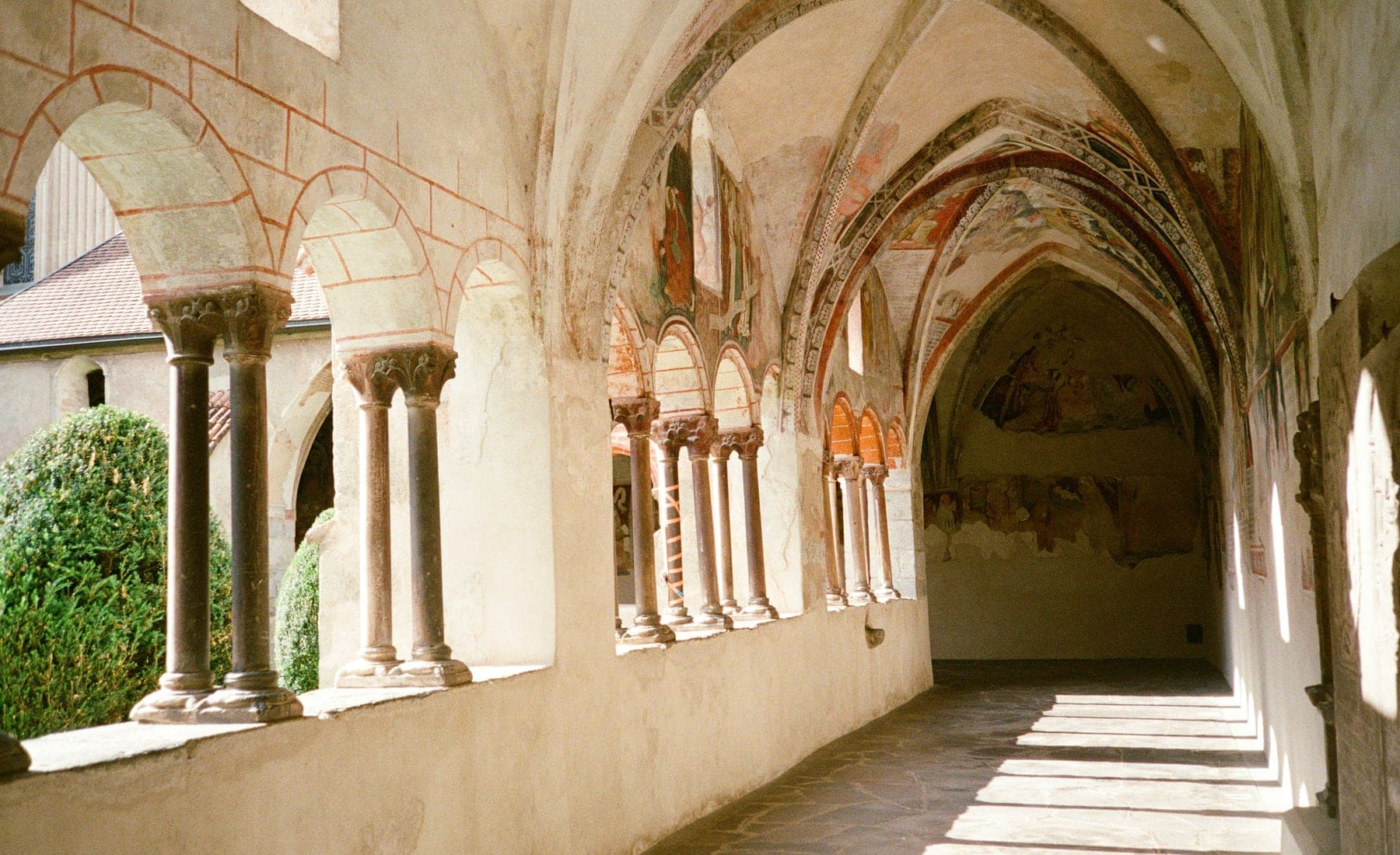 A Romanesque cloister: paired columns with carved capitals carrying round arches, faded wall paintings visible in the gallery behind.