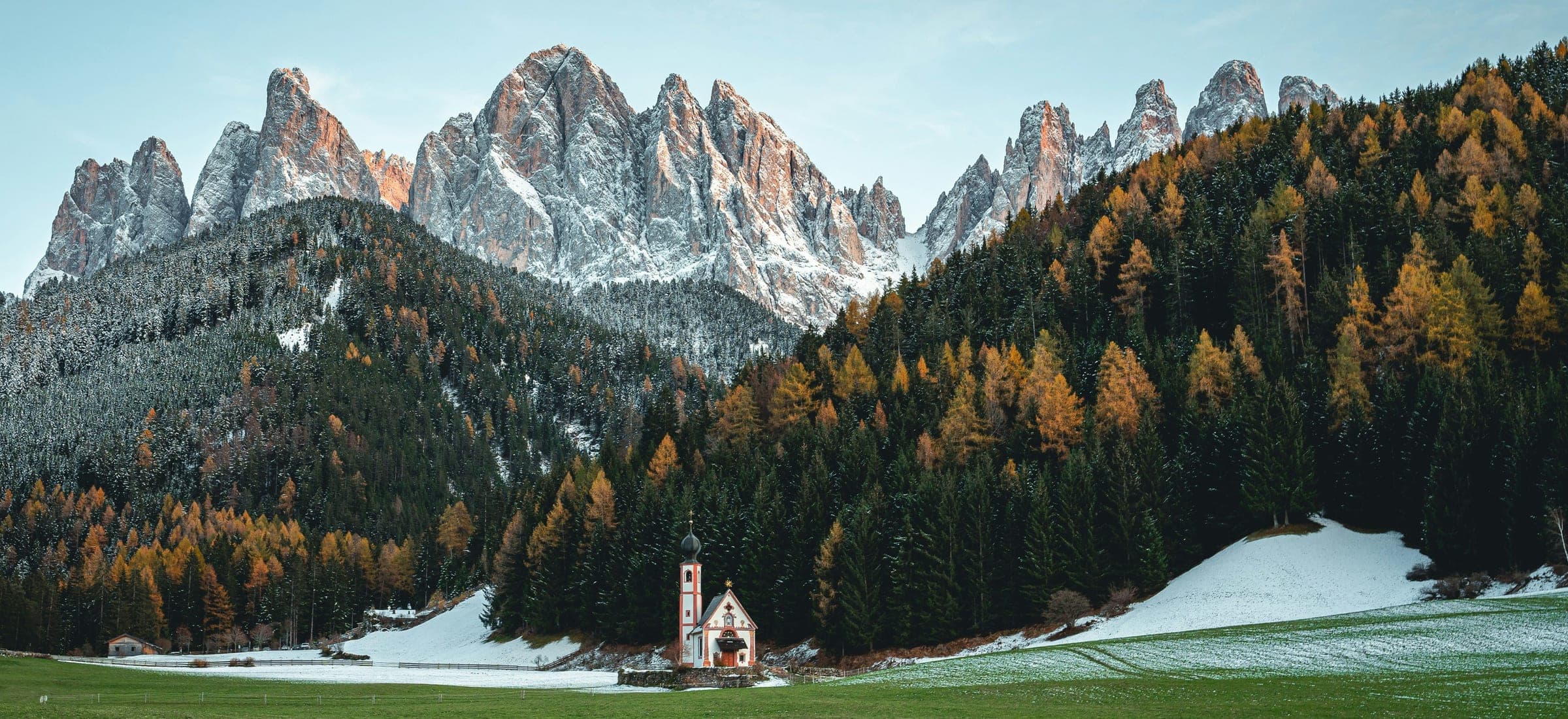 A small white chapel alone in a mown meadow, below wooded slopes turning for autumn and a ridge of bare rock peaks under fresh snow.