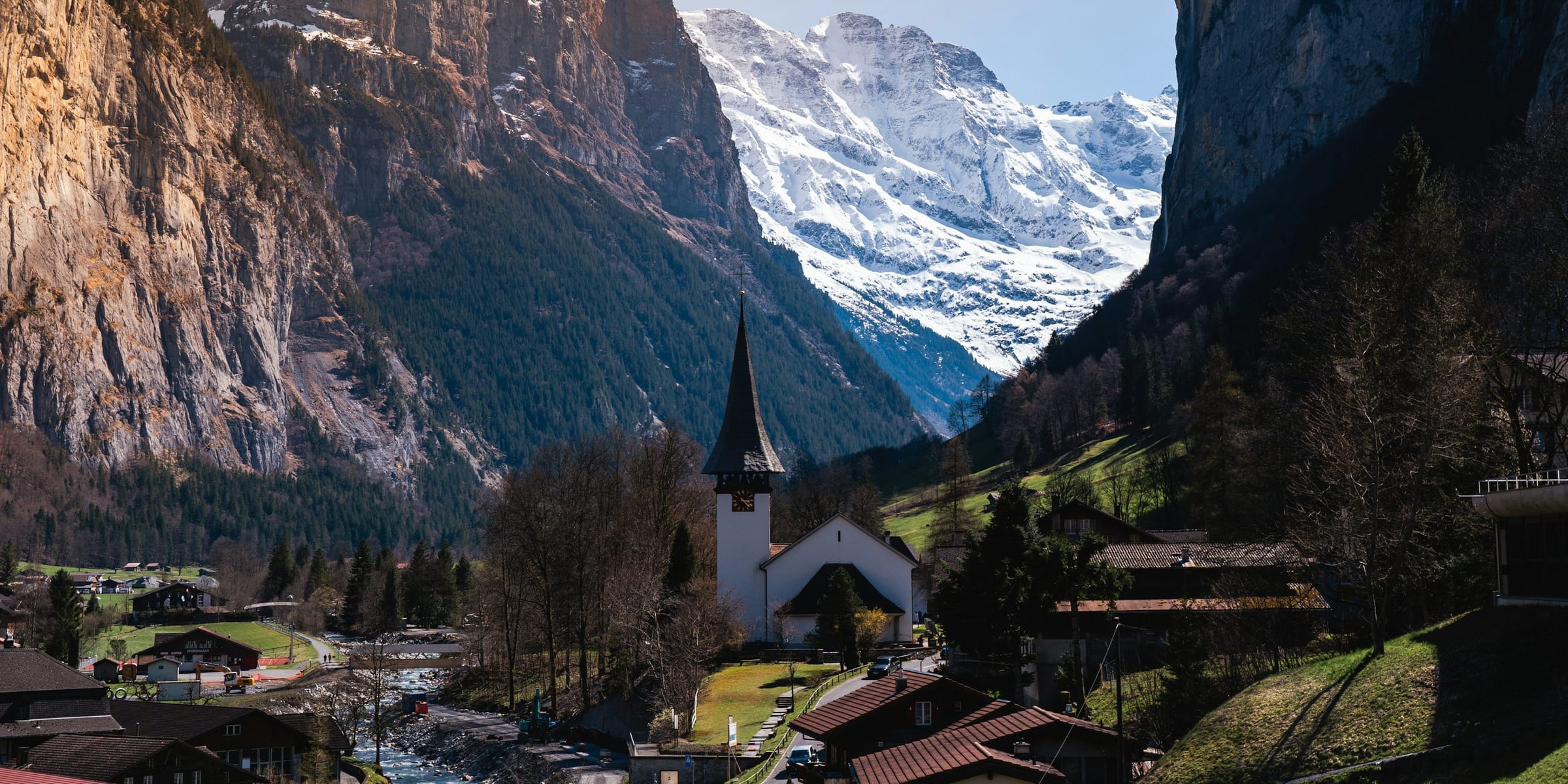 A village gathered around its church on the floor of a steep alpine valley, sheer cliffs on either side and a snow-covered massif closing the head of the valley.